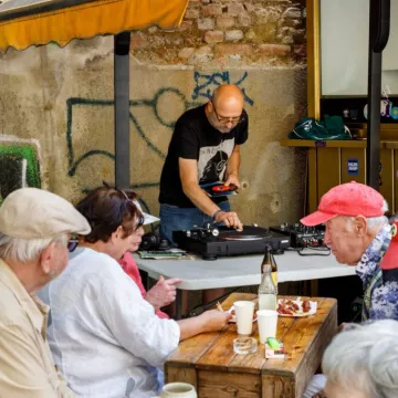 Mehrere Personen sitzen, im Hintergrund steht eine Person vor Plattenspielern und Mischpult. 
