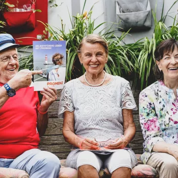 Drei Personen sitzen nebeneinander, eine Person hält eine Broschüre hoch auf der steht: Selbstständig leben mit PROMENZ. 