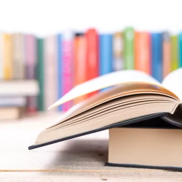 Ein aufgeschlagenes Buch liegt über einem anderen. Im Hintergrund befinden sich verschwommen aufgestellte Bücher.