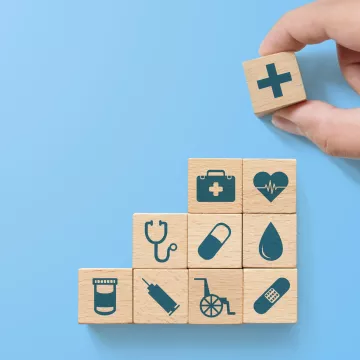 Holzblöcke mit medizinischen Symbolen (z. B. Stethoskop, Herz, Spritze, Pille) sind stufenförmig aufgebaut. Eine Hand setzt oben einen Würfel mit Kreuzzeichen – Sinnbild für Gesundheitsvorsorge und Systemaufbau.