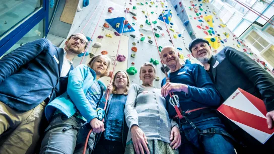 Mehrere Personen stehen vor einer Kletterwand und schauen in die Kamera. 