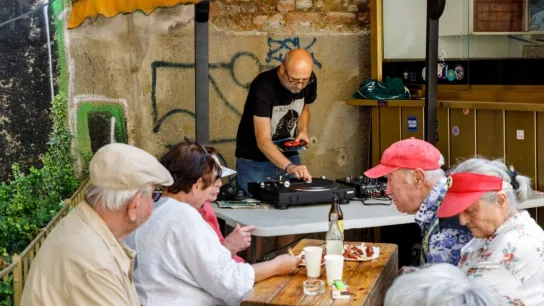 Mehrere Personen sitzen, im Hintergrund steht eine Person vor Plattenspielern und Mischpult. 