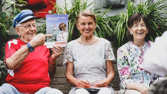 Drei Personen sitzen nebeneinander, eine Person hält eine Broschüre hoch auf der steht: Selbstständig leben mit PROMENZ. 