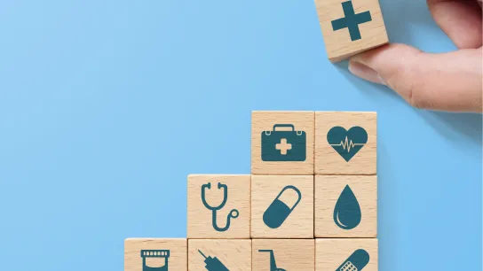 © Monster Ztudio - stock.adobe.com Holzblöcke mit medizinischen Symbolen (z. B. Stethoskop, Herz, Spritze, Pille) sind stufenförmig aufgebaut. Eine Hand setzt oben einen Würfel mit Kreuzzeichen – Sinnbild für Gesundheitsvorsorge und Systemaufbau.